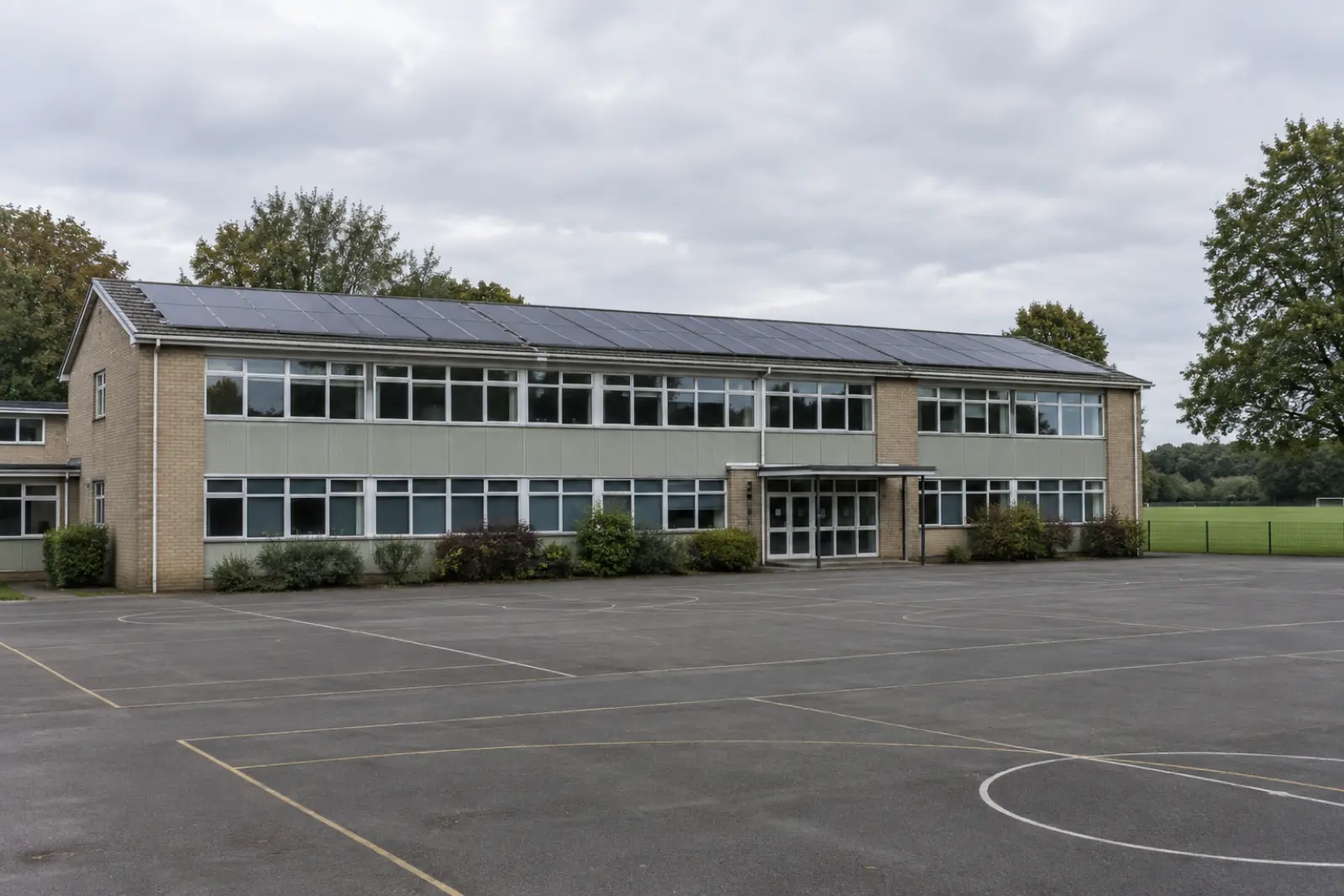 A two storey 1970s school block with a shallow pitched roof covered in solar panels, empty playground in front and a playing field beyond